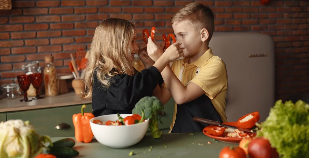 Playful boy and girl wearing apron preparing healthy vegetarian salad together at table with ingredients and bowls while looking at each other through slices of pepper